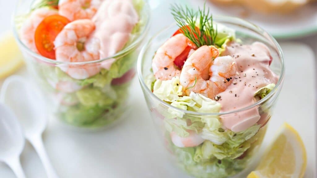 Two glass cups filled with shrimp cocktail, layered with lettuce, cherry tomatoes, and creamy pink sauce, garnished with dill. A classic 80s food fad appetizer.