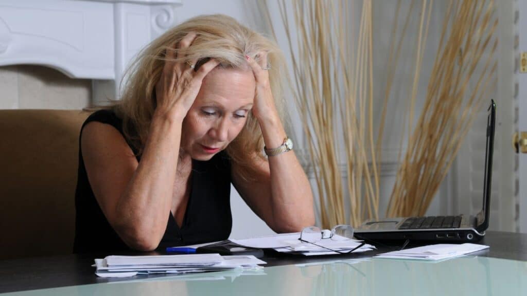 A stressed older woman sits at a desk with her hands in her hair, surrounded by bills and a laptop, conveying frustration often linked to brain fog.