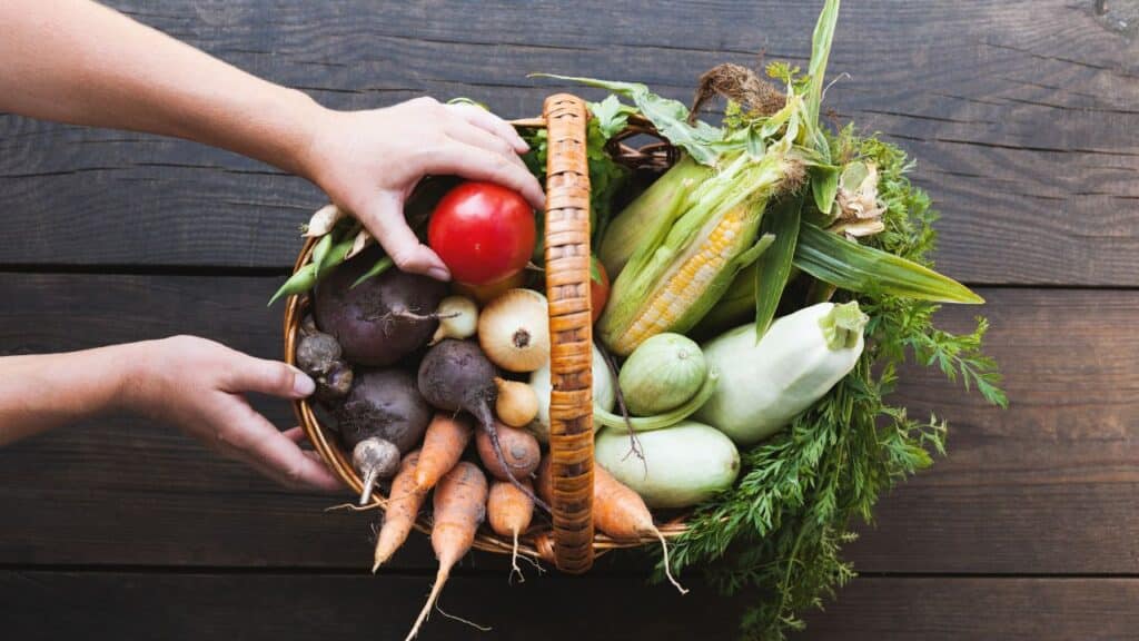 Hands arranging fresh vegetables like carrots, corn, onions, and tomatoes in a basket, emphasizing the nutritional benefits of seasonal foods in a healthy diet.