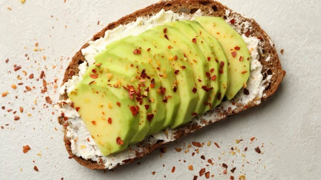 Sliced avocado on whole-grain toast with cream cheese and red pepper flakes sprinkled on top. A nutritious snack rich in healthy fats and considered one of the best anti-aging foods.