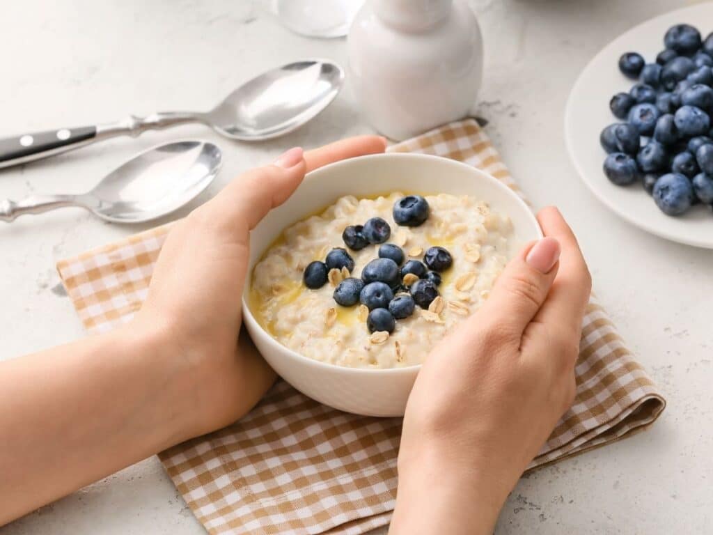Hands holding a white bowl of oatmeal topped with fresh blueberries on a checkered napkin. A plate of extra blueberries and two spoons sit nearby, highlighting this healthy anti-aging food combination.