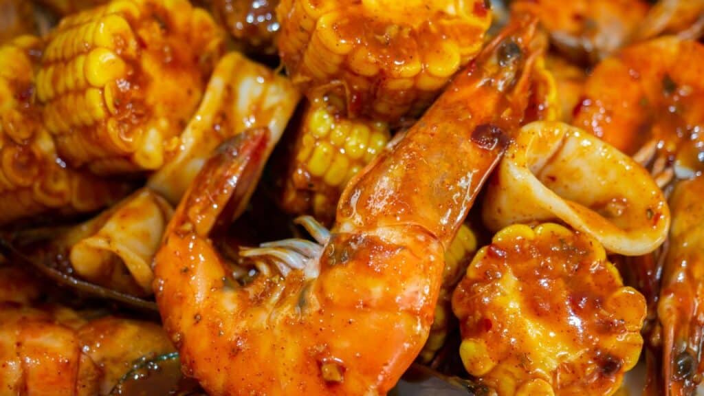 Close-up of a spicy seafood boil with shrimp, corn on the cob, and shellfish coated in a rich, seasoned sauce. A hearty 80s food fad popular at gatherings.