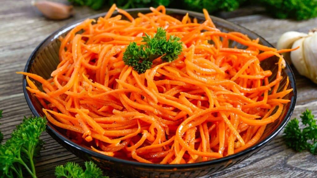 A bowl of shredded carrot salad garnished with parsley on a rustic wooden table. Carrots are full of beta-carotene and make a great anti-aging food.
