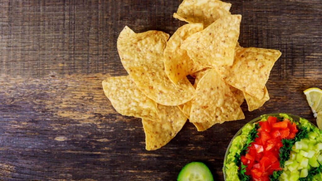Crispy tortilla chips served next to fresh guacamole topped with diced red peppers, green onions, and cucumber slices on a rustic wooden table. A vibrant 80s food fad snack.
