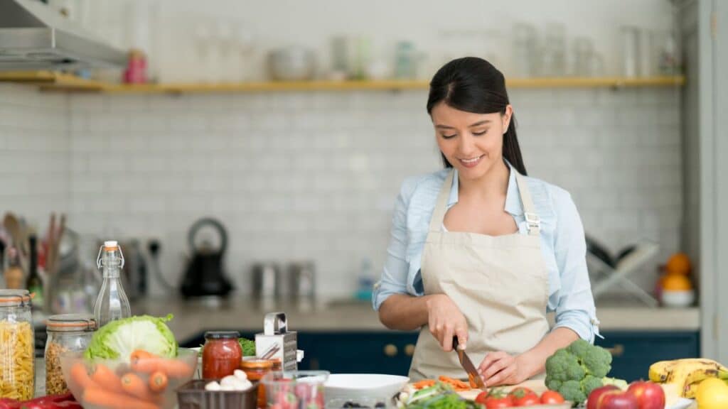 Woman in a bright kitchen chopping fresh vegetables like carrots, broccoli, and tomatoes, demonstrating the nutritional benefits of seasonal foods in home cooking.