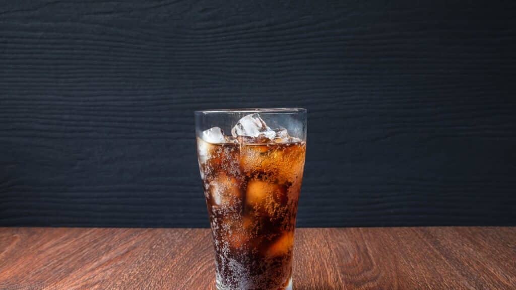 A tall glass of cola with ice cubes on a wooden table against a dark background, highlighting the fizzy refreshment. A staple drink during 80s food fads.
