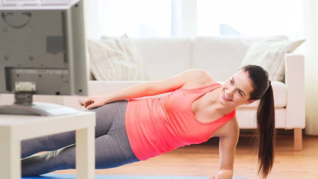Woman exercising at home on a yoga mat in front of a TV, smiling during a workout to hit 10000 steps a day.