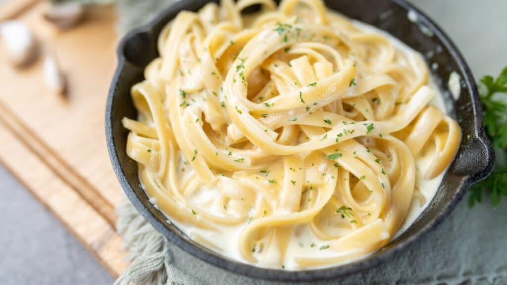 Creamy fettuccine Alfredo pasta in a black bowl, sprinkled with herbs, showcasing the rich sauce and tender noodles. A beloved 80s food fad dish.
