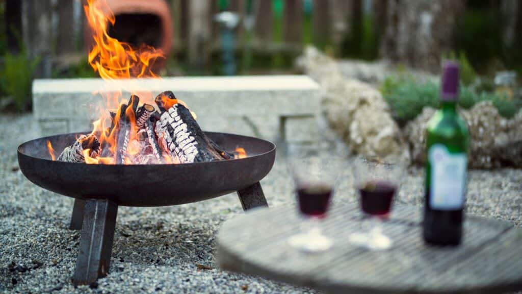 A crackling fire pit with glowing embers next to a table holding two glasses of red wine, creating a warm and inviting outdoor atmosphere.