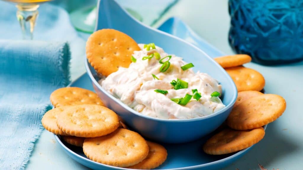 Round golden crackers arranged around a creamy dip topped with green onions in a blue dish, served on a pastel table setting. A go-to 80s food fad party snack.