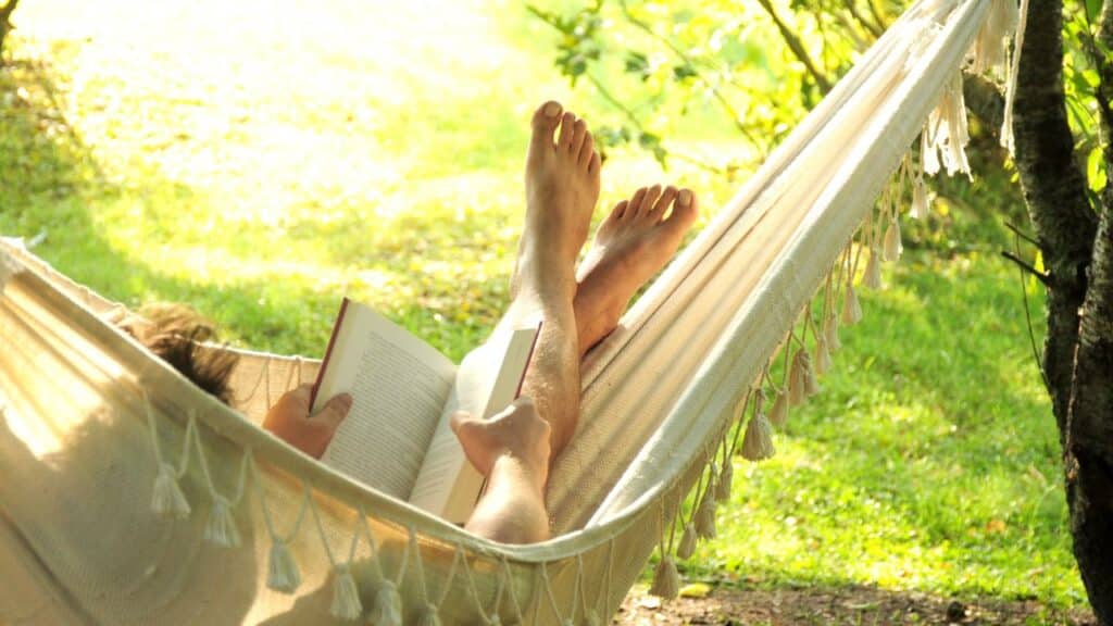 Person relaxing in a hammock under tree shade while reading a book, embodying the perfect way to turn your backyard into a relaxing haven.