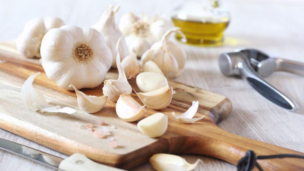 Whole garlic bulbs and peeled cloves on a wooden cutting board with a garlic press in the background. Garlic is known for its health benefits and is considered an anti-aging food.
