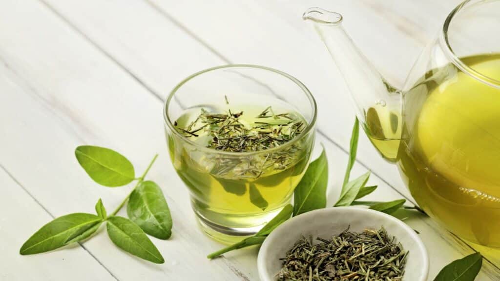 A glass of green tea with fresh leaves and a teapot in the background on a white wooden table. Green tea is a powerful anti-aging food rich in antioxidants.