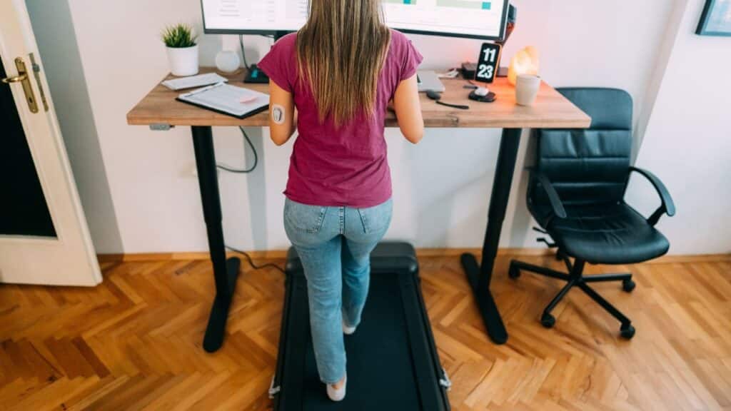 Woman using a treadmill under a standing desk while working on a computer, multitasking to hit 10000 steps a day.