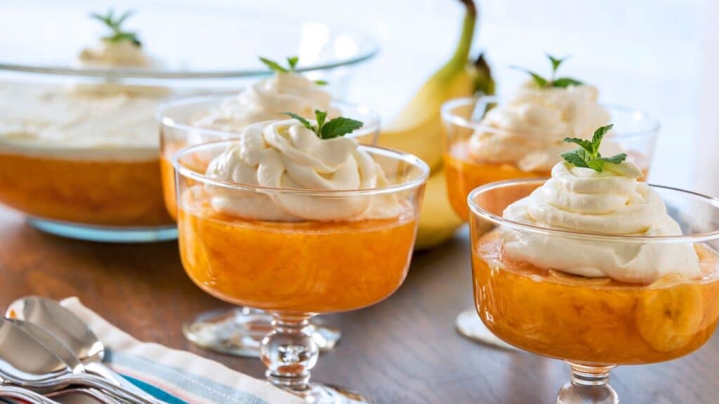 Glass dessert cups filled with bright orange banana pudding topped with whipped cream and mint leaves, with bananas in the background. A sweet 80s food fad dessert.