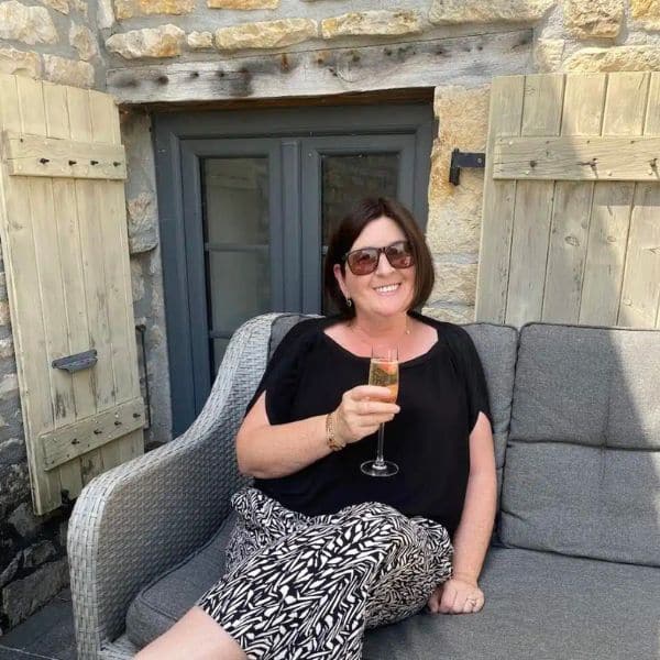 A middle aged woman holding a glass of champagne sat on grey couch outside with sun glasses on and dark hair.