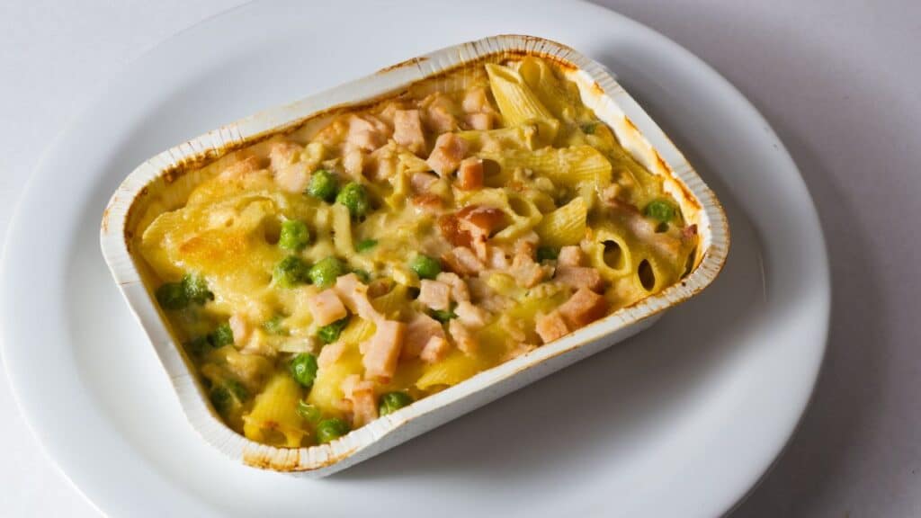 A baked chicken tetrazzini casserole in a white foil container with penne pasta, peas, and creamy sauce, slightly browned on top. A comforting 80s food fad meal.