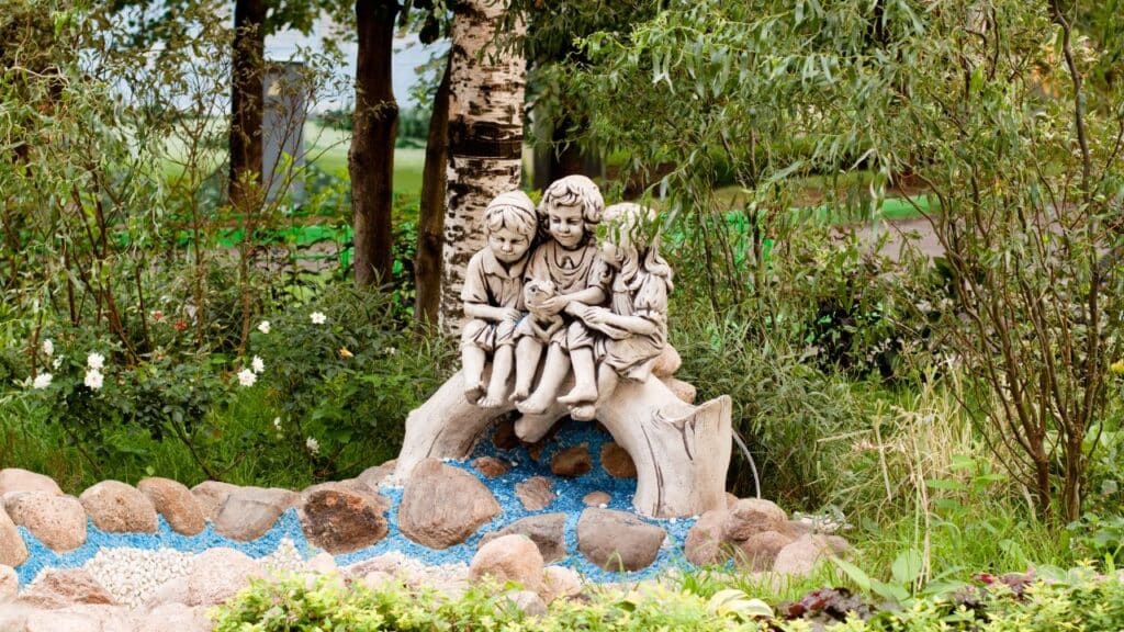A stone sculpture of three children sitting on a log with a bird, nestled in a lush garden, bringing a playful yet tranquil vibe.