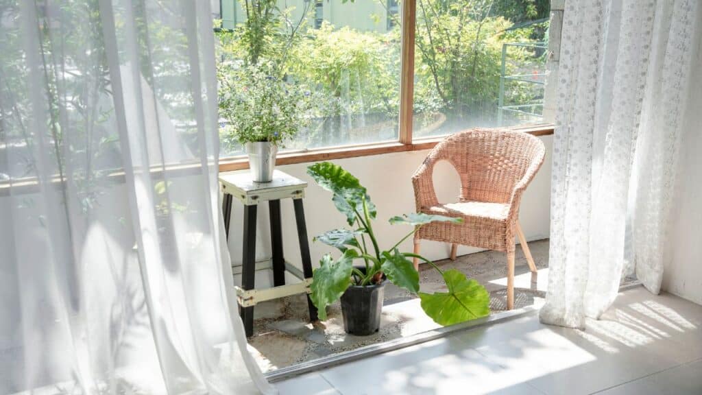 A sunlit indoor-outdoor nook with wicker chair, potted plants, and sheer white curtains flowing in the breeze for a cozy, relaxing vibe.