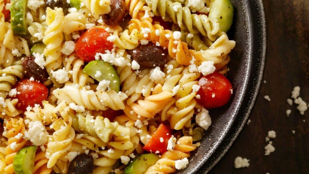 Colorful pasta salad with rotini, cherry tomatoes, black olives, cucumber slices, and crumbled feta on a black plate. A refreshing 80s food fad potluck favorite.