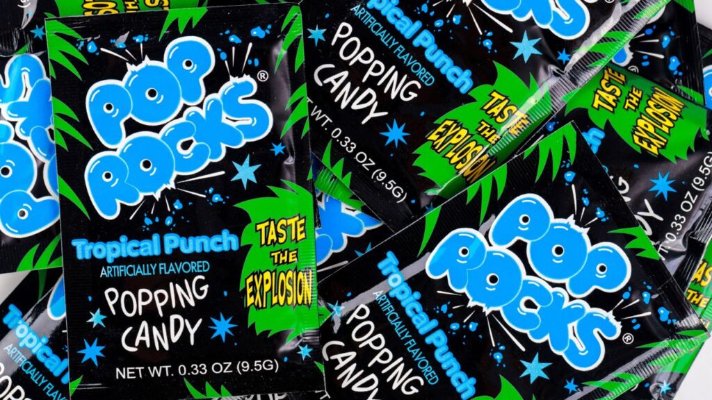 Several packets of Pop Rocks Tropical Punch popping candy with bold blue and green graphics, featuring the phrase “Taste the Explosion.” A nostalgic 80s food fad candy.