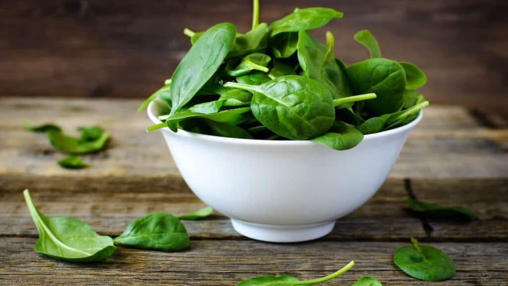 Fresh spinach leaves in a white bowl on a wooden surface, with a few leaves scattered around. Spinach is packed with nutrients and is a classic anti-aging food.