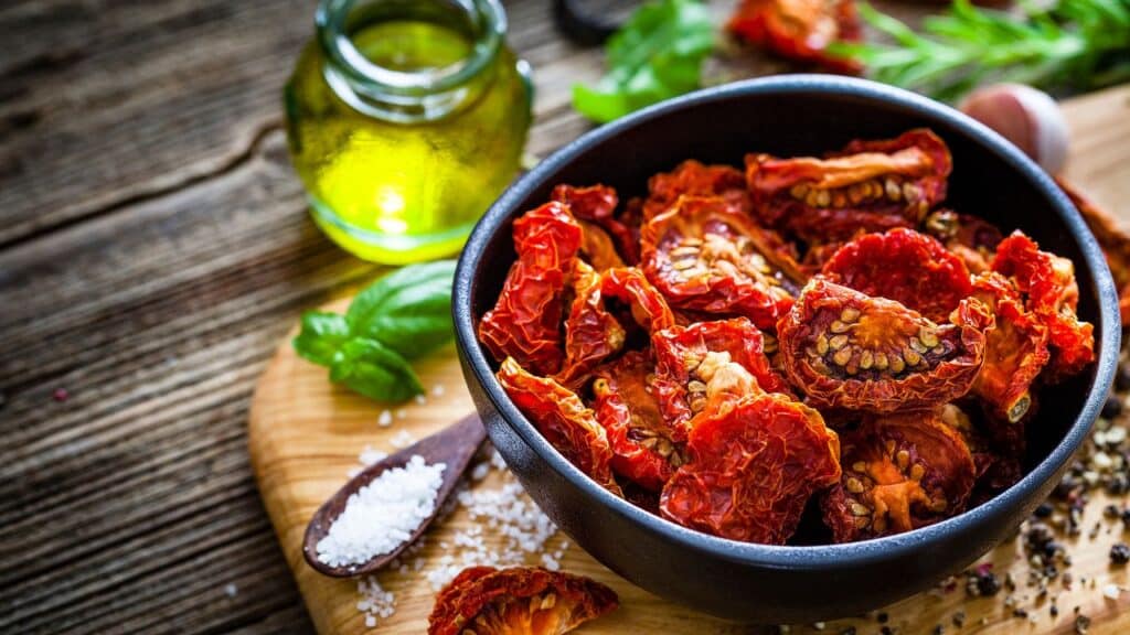 A black bowl filled with vibrant red sun-dried tomatoes on a wooden board, next to coarse salt and olive oil. A gourmet ingredient that became a major 80s food fad.