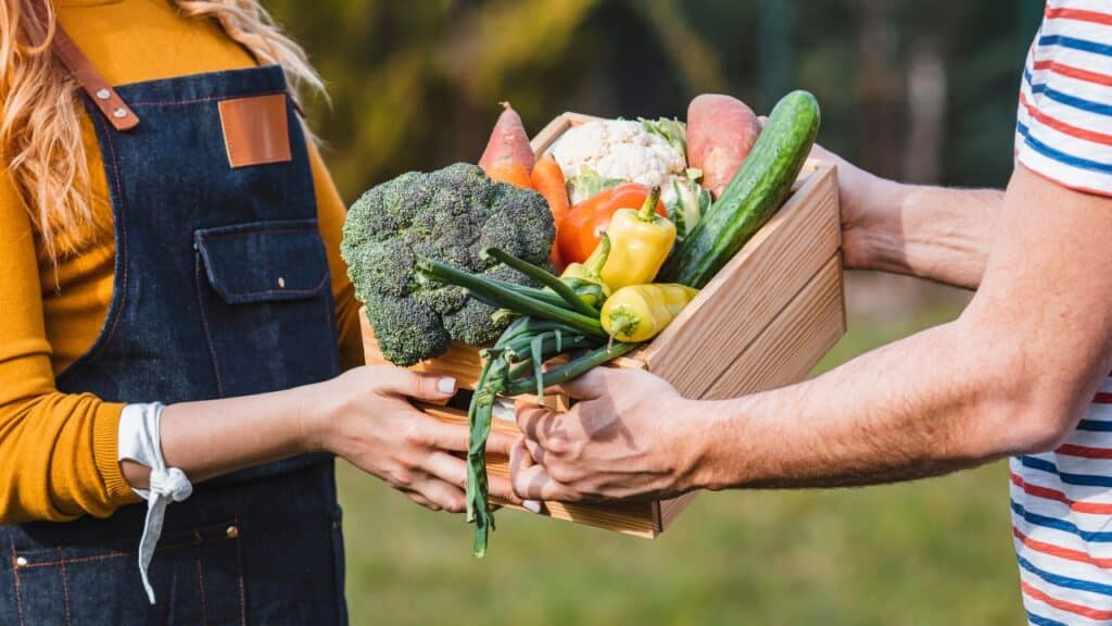 wo people exchanging a wooden box filled with broccoli, peppers, cucumbers, and other vegetables, emphasizing the nutritional benefits of seasonal foods through farm-to-table sharing.