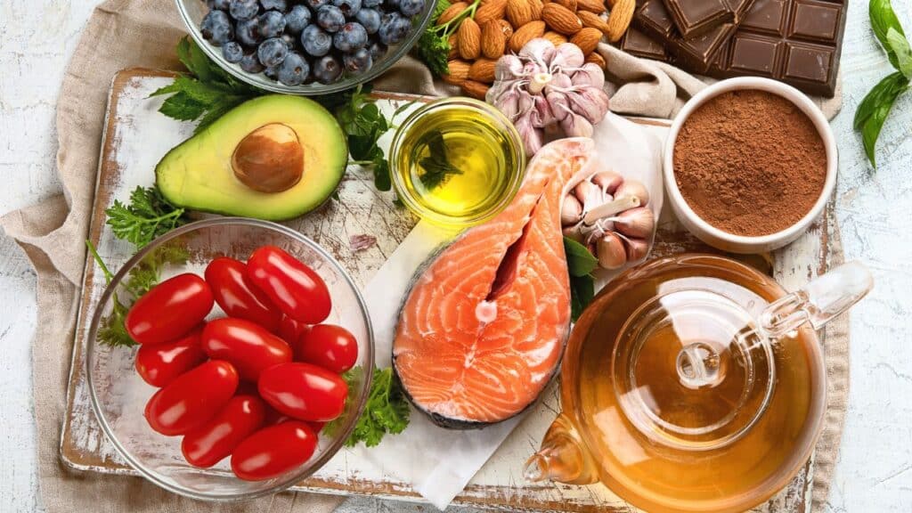 A variety of anti-aging foods including salmon, avocado, olive oil, blueberries, almonds, garlic, dark chocolate, tomatoes, green tea, and cocoa powder arranged on a rustic tray.