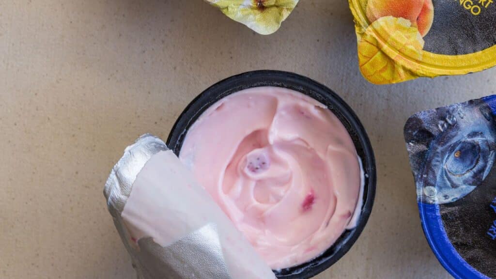 Open container of pink strawberry yogurt with foil lid peeled back, surrounded by other fruit-flavored yogurt cups. Flavored yogurt was a popular 80s food fad for health-conscious eaters.