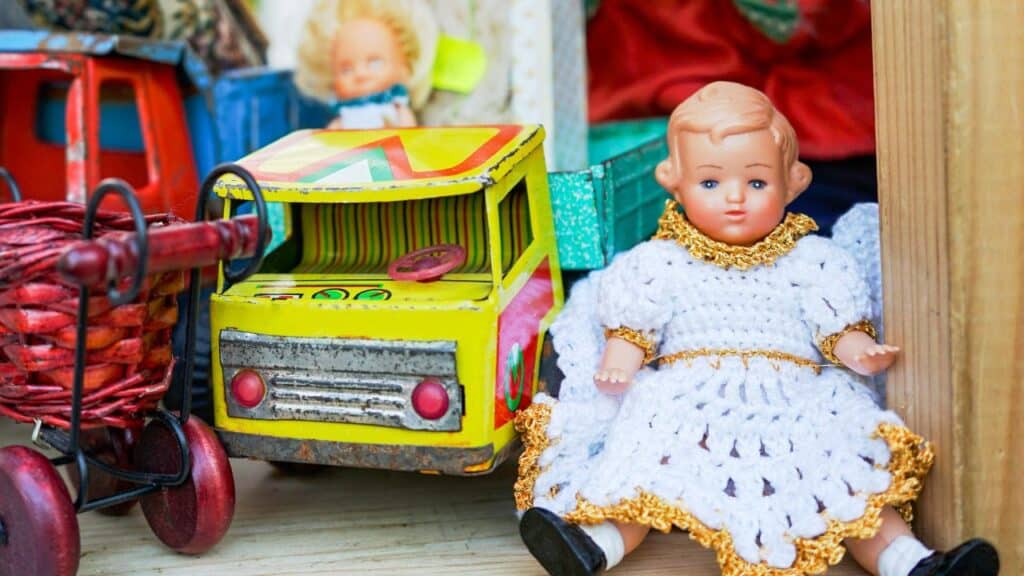 A vintage baby doll in a crocheted white dress with gold trim sits beside a colorful metal toy truck and a red wicker toy cart. These nostalgic items reflect popular childhood toys from the 70s.