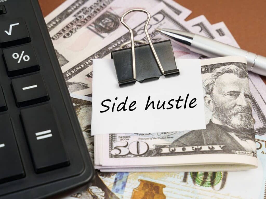 A stack of fifty-dollar bills held together with a binder clip sits on a desk next to a calculator and pen, with a note labeled “Side hustle.” Represents financial planning or earnings from smart side hustles.