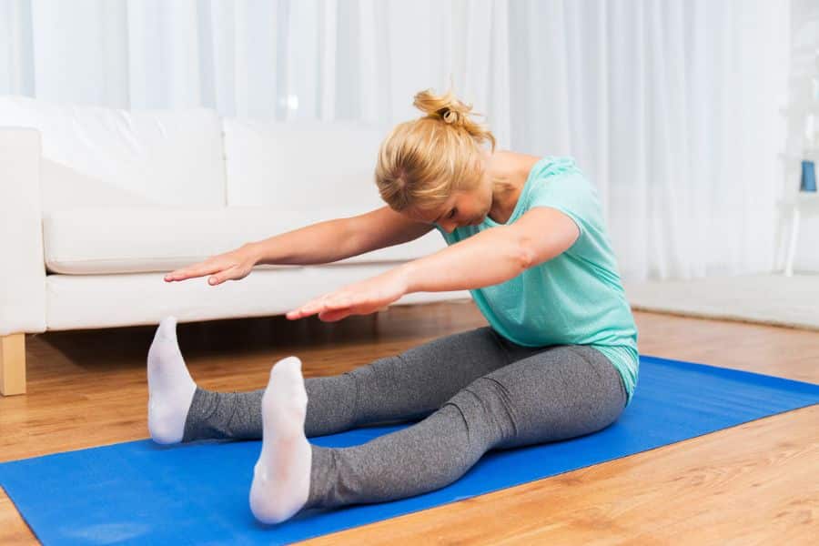 A woman in workout clothes seated on a yoga mat doing a forward stretch with both arms extended toward her feet. Gentle nighttime stretches like this can be part of nightly rituals that rewind the clock on aging.