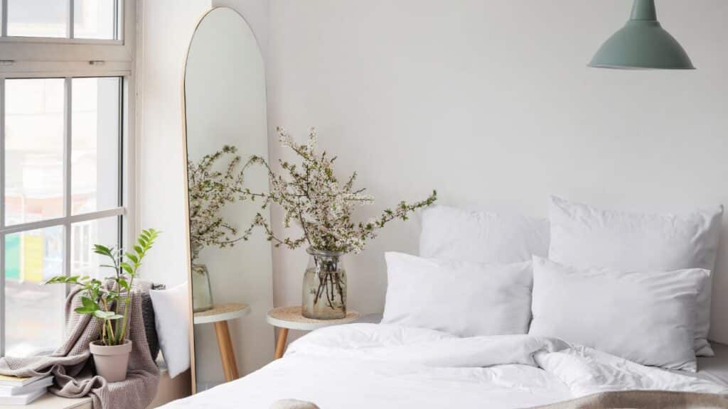 Cozy bedroom with white bedding, large mirror, and fresh flowers to attract positive chi as suggested by Feng Shui tips.