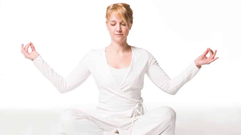 A woman in white clothes sits cross-legged on the floor with eyes closed, meditating in a bright, white space.