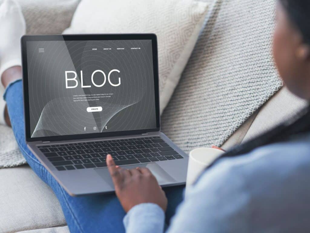 A person sitting on a couch holds a coffee mug while using a laptop that displays a minimalist blog homepage. Suggests blogging as one of many smart side hustles done from home.