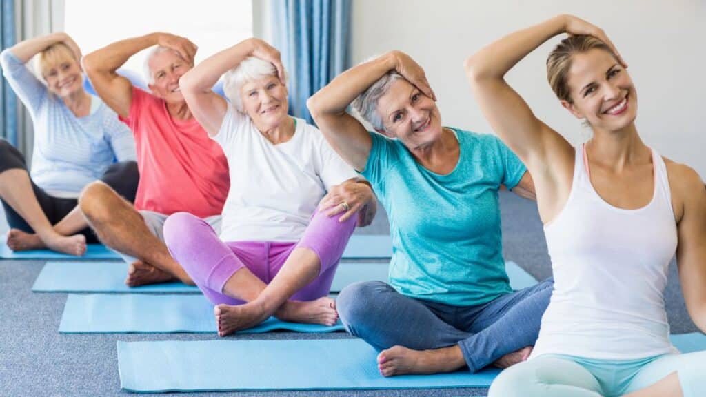 A group of older adults in colorful workout clothes sits on yoga mats indoors, tilting their heads to the side while smiling.