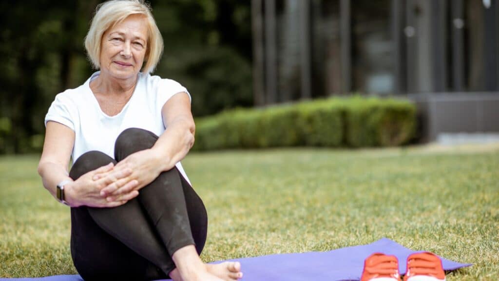 An older woman sits on a purple yoga mat outside on green grass, hugging her knees with a relaxed expression.
