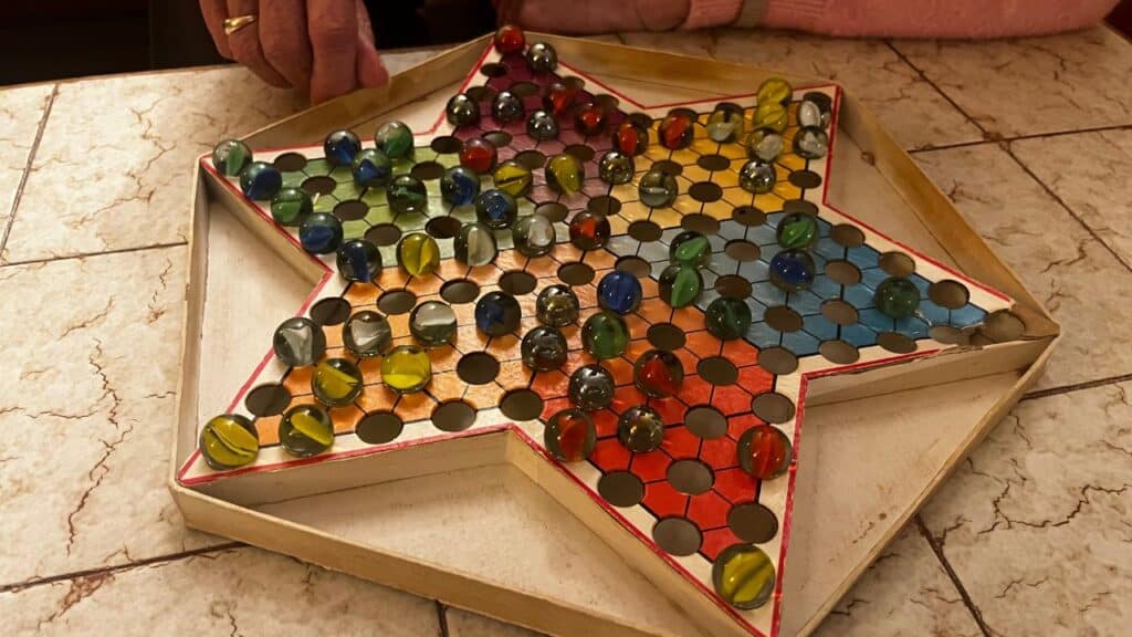 A hand reaches toward a colorful Chinese Checkers board filled with glass marbles on a star-shaped wooden board. This classic board game is a familiar part of childhood toys from the 70s.