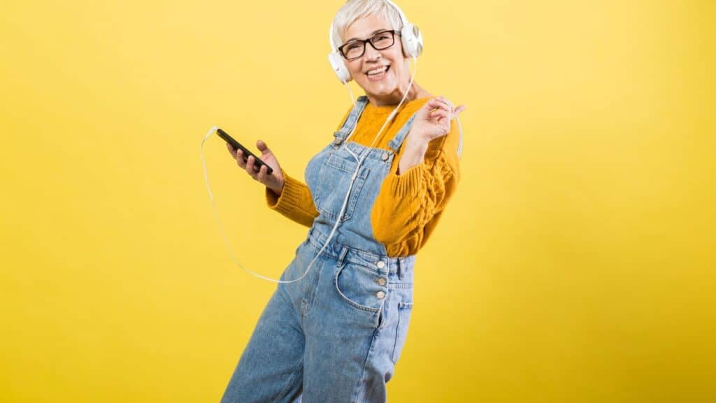 Smiling woman in denim overalls and mustard sweater listening to music on headphones, illustrating a casual outfit that might border on fashion faux pas.
