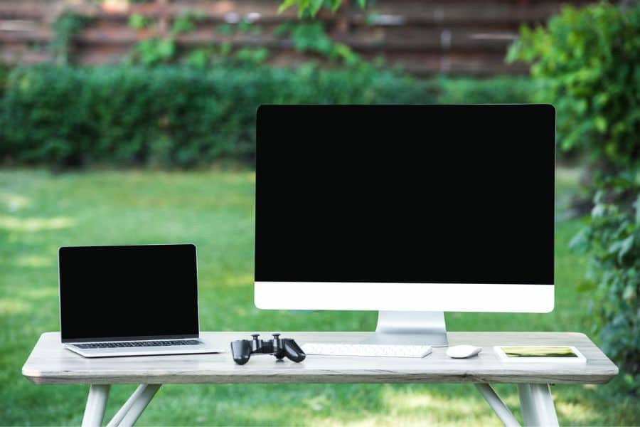 A desktop computer and a laptop with blank screens are set up on an outdoor table with game controllers and a smartphone, suggesting used electronics for sale on Facebook Marketplace.