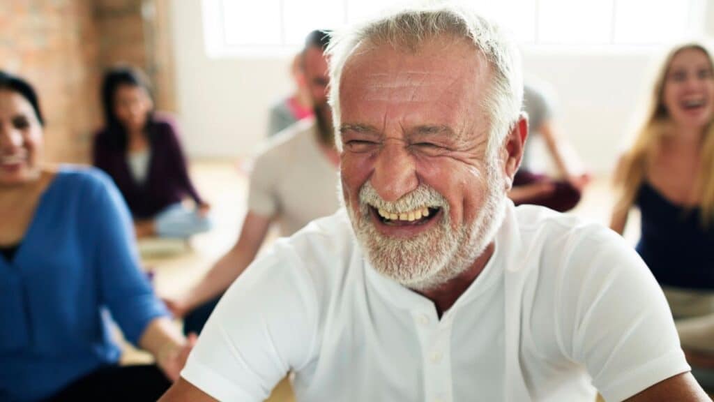 A man with a white beard and white shirt laughs joyfully during a group yoga class, with other smiling participants blurred in the background.