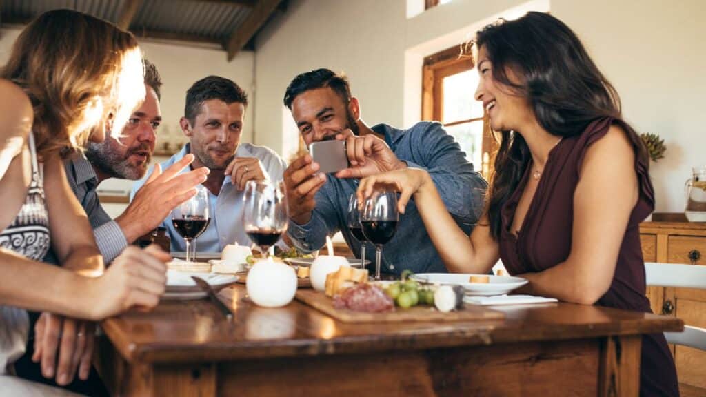 Group of friends at a summer dinner party laughing and looking at a phone while enjoying wine and appetizers.