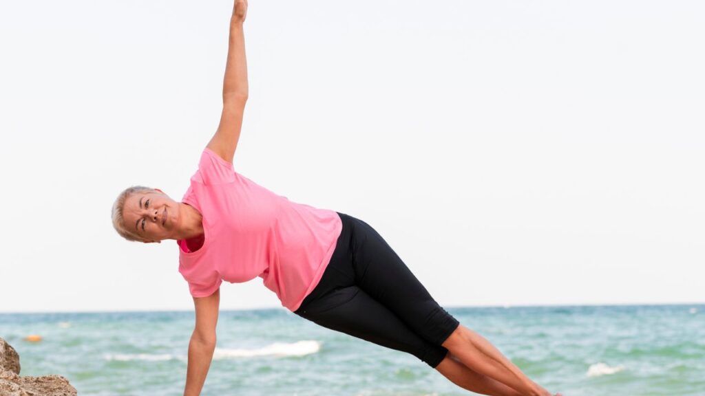 An older woman in a pink top and black shorts performs a side plank on rocks near the ocean with waves in the background.