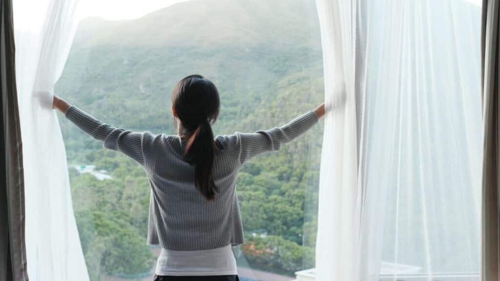 Woman opening sheer curtains to let natural light into a room, a key Feng Shui tip for inviting good energy.