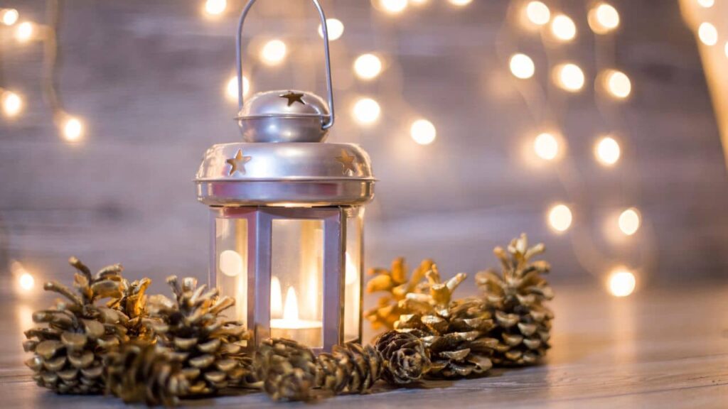 A silver lantern with candlelight within and pinecones surrounding it.