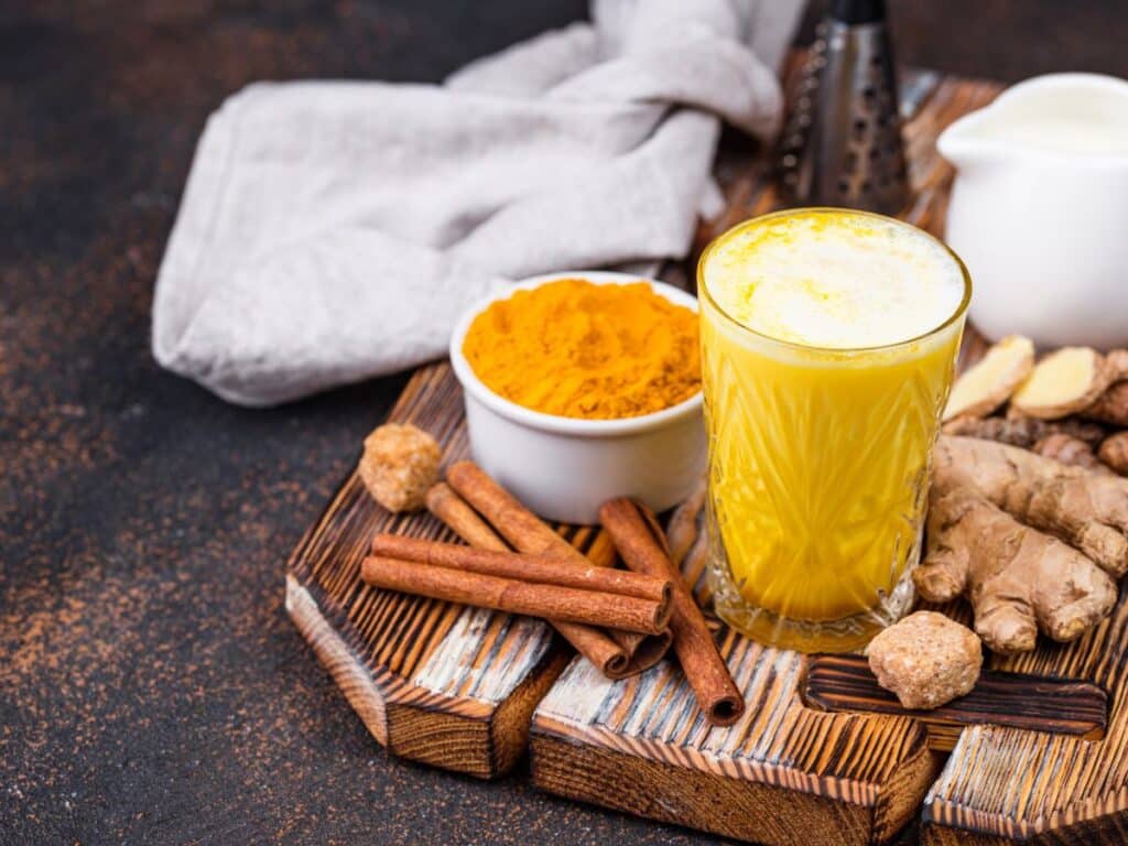 Glass of golden turmeric milk surrounded by fresh ginger, cinnamon sticks, brown sugar cubes, and a bowl of turmeric powder on a rustic wooden tray. Known for its anti-inflammatory properties, this warm drink is one of the natural anti-aging secrets rooted in Ayurvedic tradition.