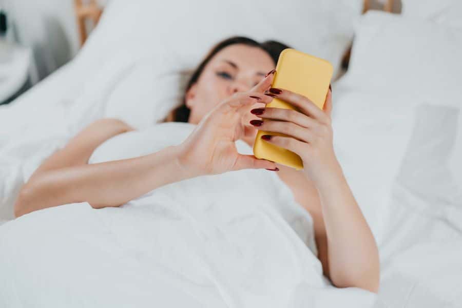 A woman lying in bed under white covers, holding a yellow smartphone above her as she scrolls. Late-night screen time contrasts with effective nightly rituals that rewind the clock on aging.