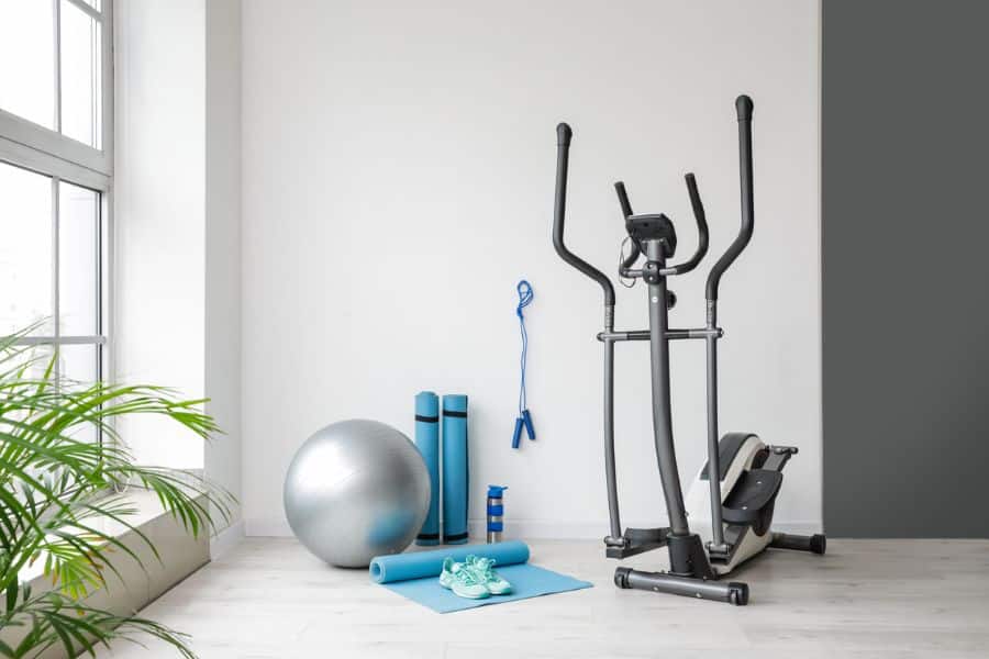 A home gym setup featuring an elliptical machine, yoga mat, dumbbells, sneakers, and a silver exercise ball, arranged neatly in a bright corner for Facebook Marketplace resale.