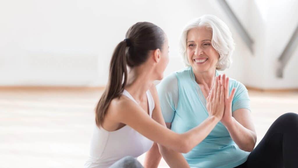 Two women sitting on the floor indoors smile and give each other a high-five after exercising.